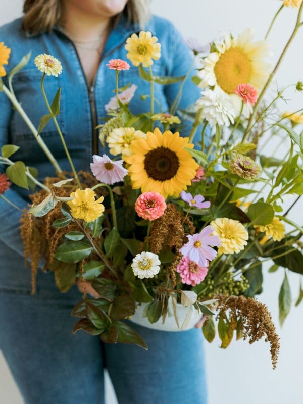 floral2 A woman holding a vase full of flowers. She is wearing blue jeans. The vase is filled with lots of different colored flowers.